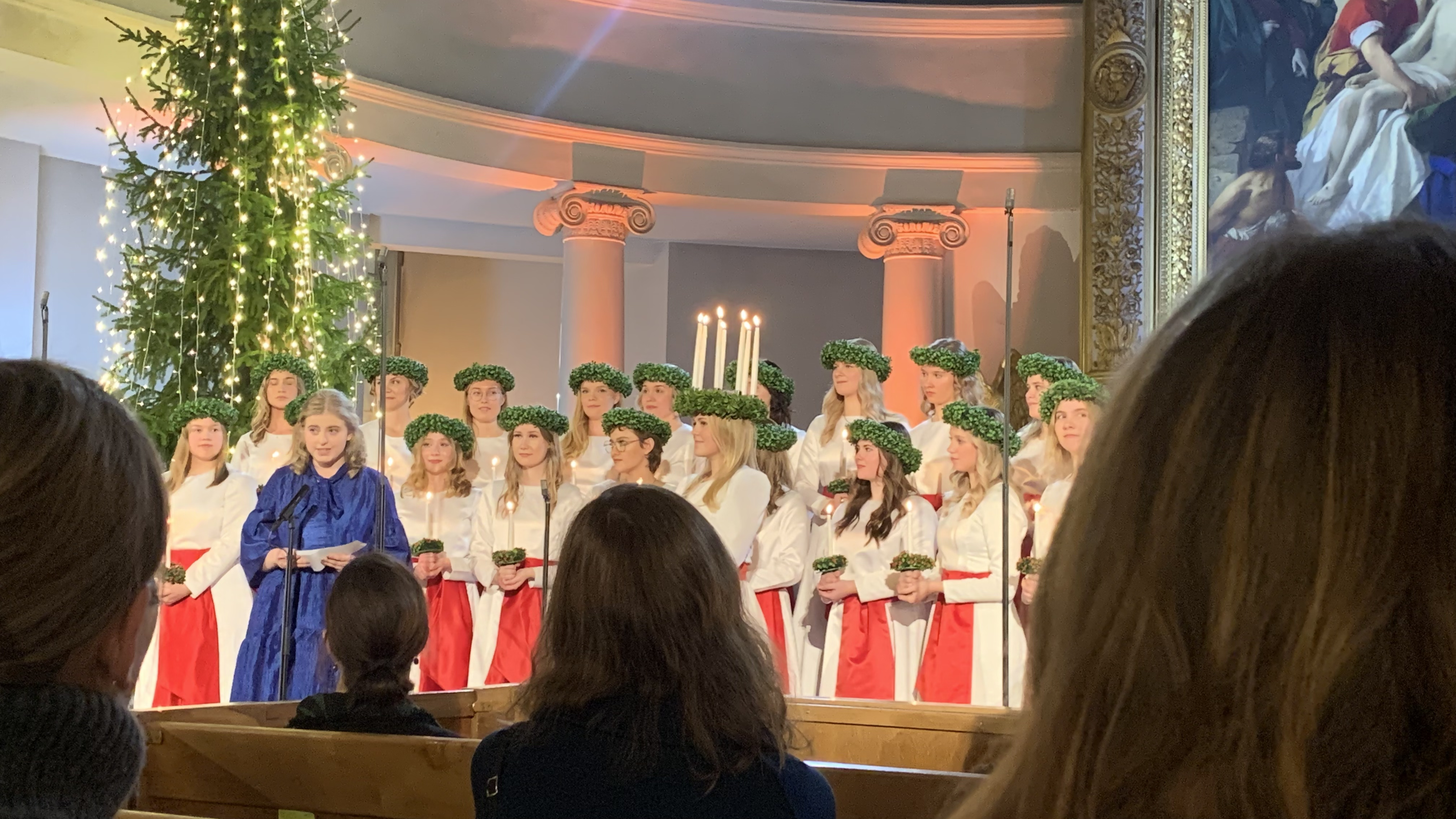 I blå klänning står FSS ordförande Cecilia Huhtala och håller tal till Finlands lucia Wilma Grönqvist i domkyrkan. Bakom lucia står luciakören i två rader.